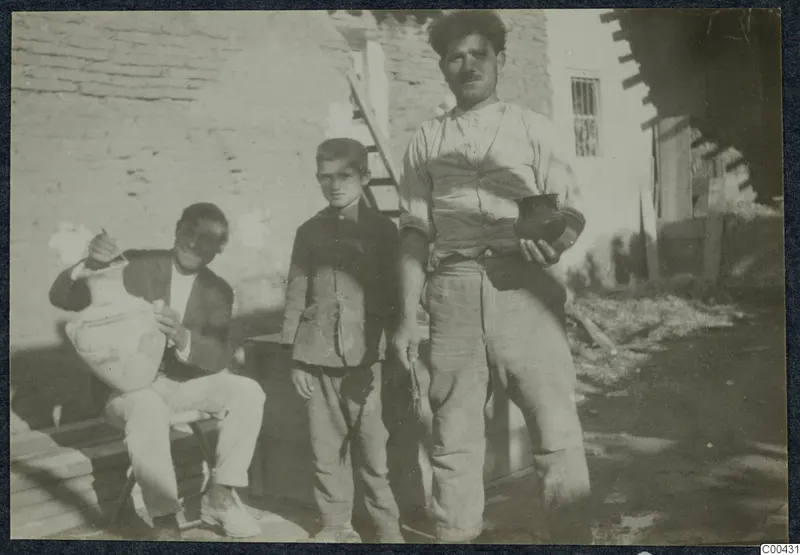 Two men and a boy outside a mud-brick wall; one seated man holds a large pale jar, and a standing man holds a small dark vessel.