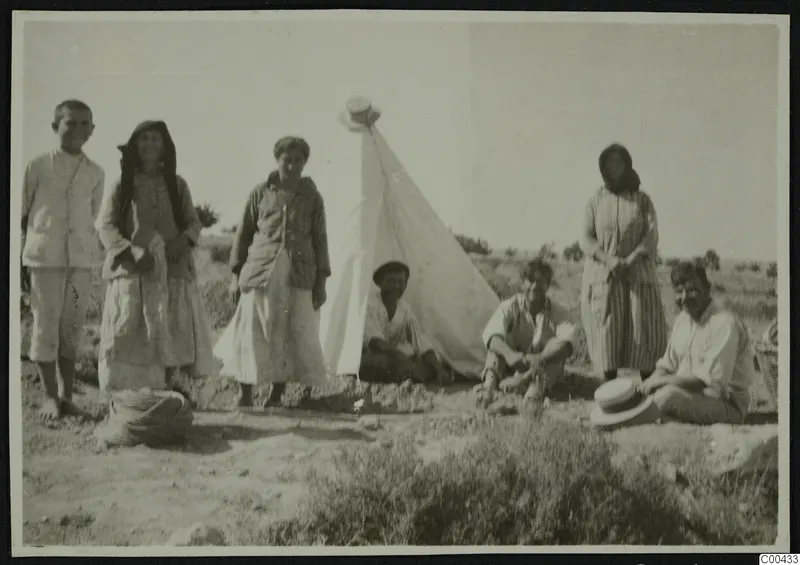 Seven people posed beside a white tent with a straw hat on its peak, on open dry ground