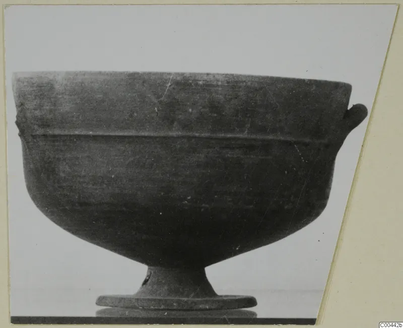 Pottery bowl on a stand, dark surface with a handle-like lug at the right rim and a footed base.