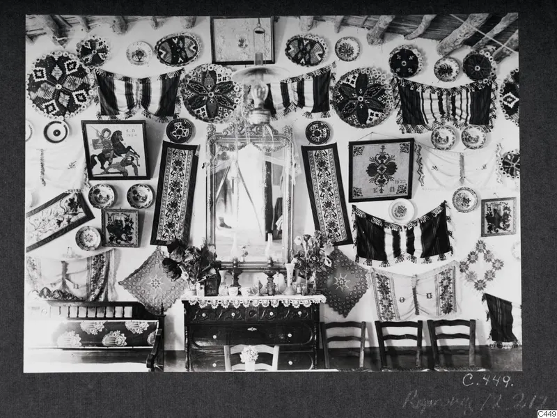 Interior of a house at Milia, a wall hung with embroidered textiles, plates, a mirror and a lamp.