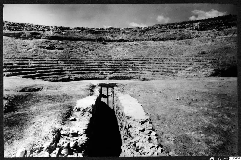 Theatre at Soli with rows of stone seating curving up a slope, beside a narrow excavated passage covered by a wooden plank.