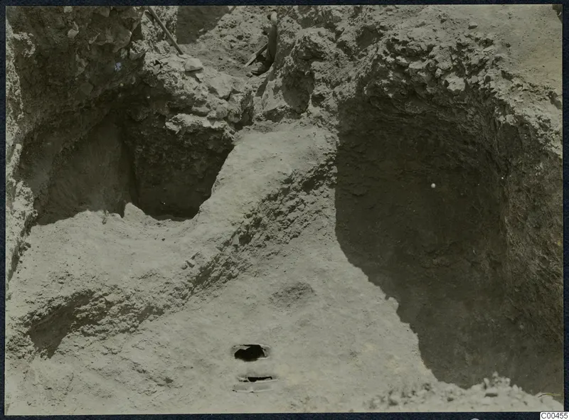 Excavation trench at Soli with cut soil faces, loose stones and a digging tool handle visible.