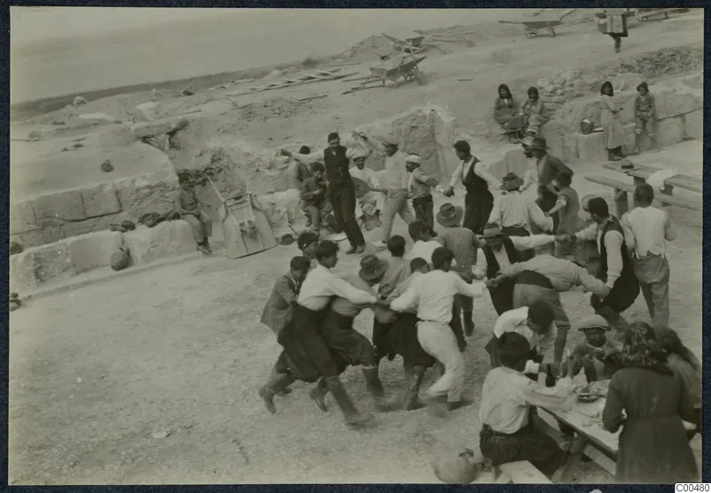 Men dancing in a linked line at the theatre at Soli, with wheelbarrows and onlookers nearby