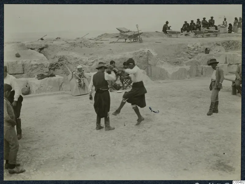 Men dancing on the stony floor of the theatre at Soli, with people sitting on wheelbarrows on the excavated ground behind.