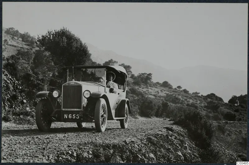 Open-top touring car with a registration plate driving along a gravel road in hills.