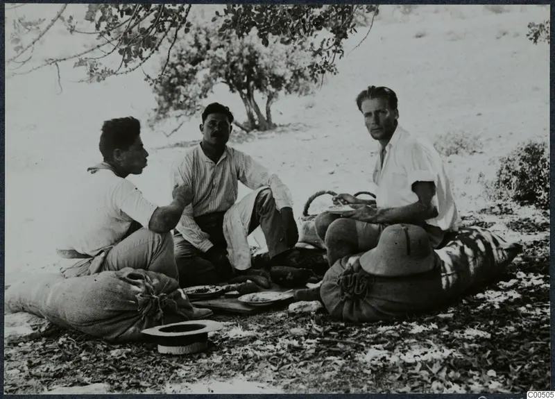 Three men eating outdoors, seated on sacks under a tree, with a pith helmet resting beside one of them.