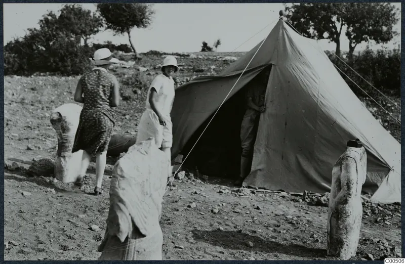 Two women and a man beside a tent at Vouni, with three stone sculptures standing outside it.