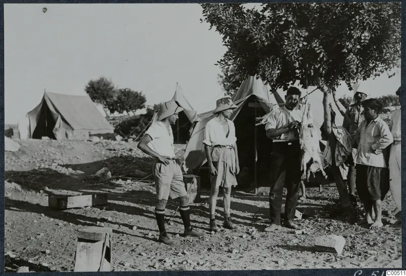 Three tents and six men standing around a tree, with an animal carcass hanging from a branch at Vouni
