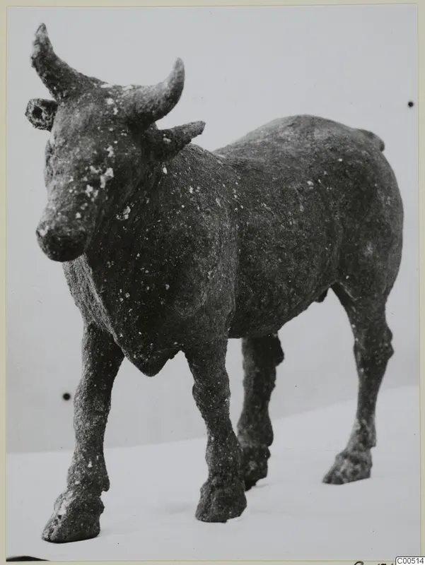 Bronze figure of a standing cow or bull with curved horns and a corroded dark surface, shown against a pale background.
