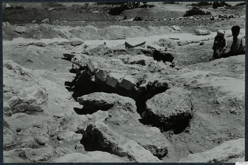 Excavation at the Temple of Athena, Vouni, with fallen stone blocks in a trench and two people seated at the right edge