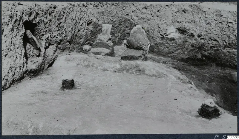 Finds in situ in an excavated trench at Vouni Palace, with stone pieces lying on a cleared floor against an earth face.