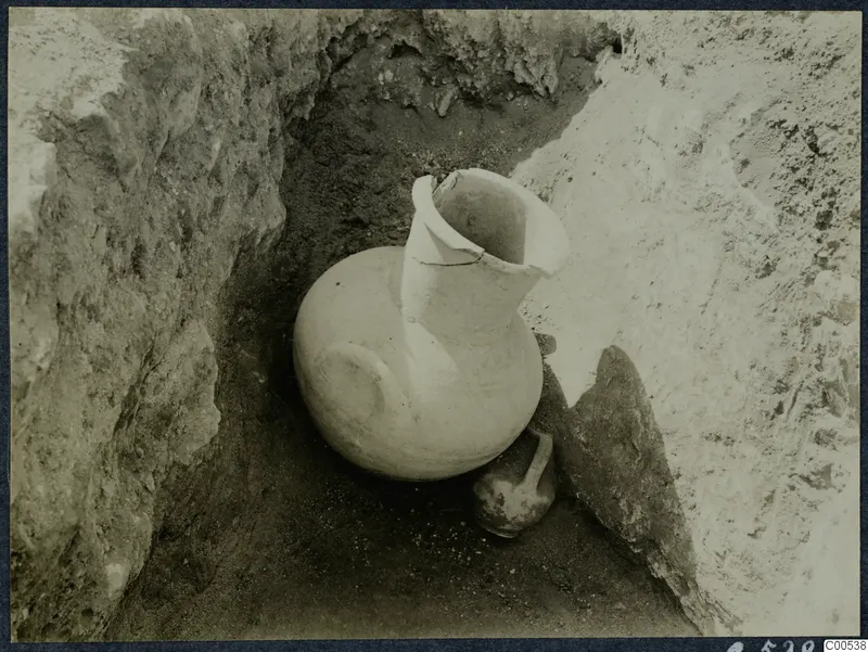 Large pale jug and a small dark vessel lying in an excavated trench at Vouni