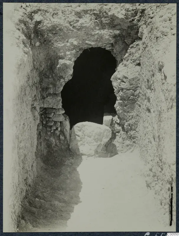 Narrow excavated passage leading to a dark arched opening, with a large pale stone block lying in front of it