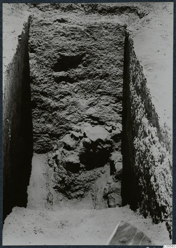 Narrow excavation trench at Vouni with a tall rough wall face and stones at its base