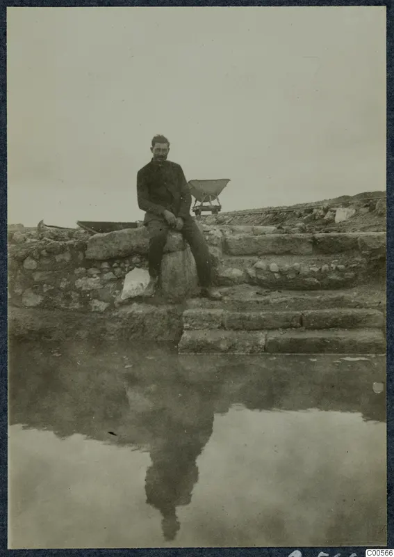 Man seated on a stone block beside stone steps and walls at Vouni, with still water in the foreground reflecting him