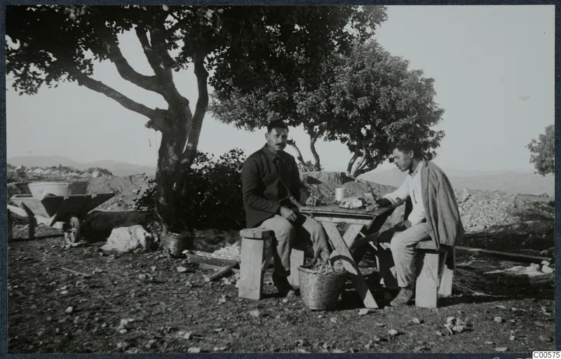 Two men seated at a wooden table under a tree at Vouni, with a wheelbarrow and baskets nearby.
