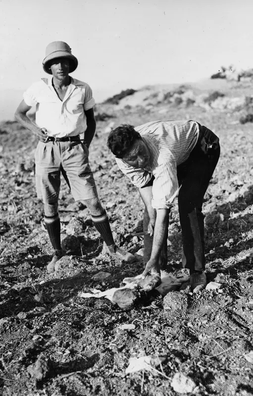 Two men with a slaughtered goat lying on stony ground at Vouni; the man on the left wears a pith helmet.