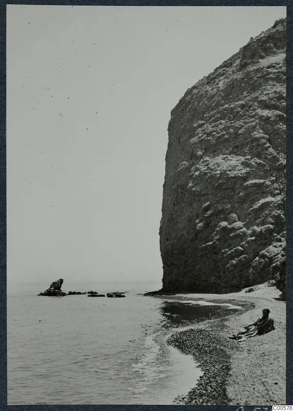 The bathing beach at Vouni, with a tall rock cliff on the right and a seated person on the pebbles.