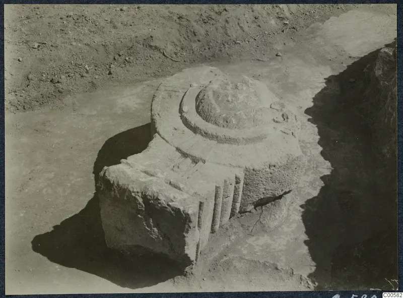 Carved stone architectural piece lying in an excavated trench at Vouni Palace, with a stepped round top and vertical grooves