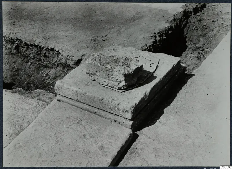 Squared stone block with a broken stone fragment on top, among slabs in an excavation at Vouni