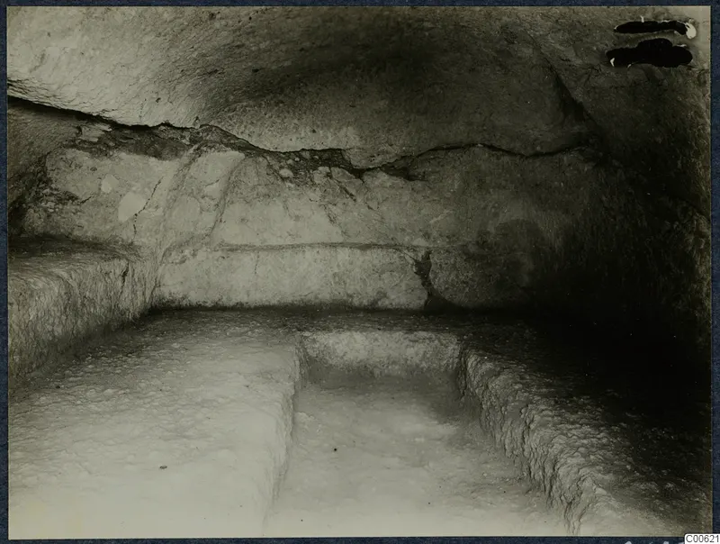 Interior of a rock-cut tomb chamber at Vouni with carved benches and a sunken rectangular area in the floor.