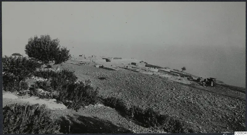 Low ruined walls and small figures on a stony hillside at Vouni, with bushes in the foreground and the sea beyond.