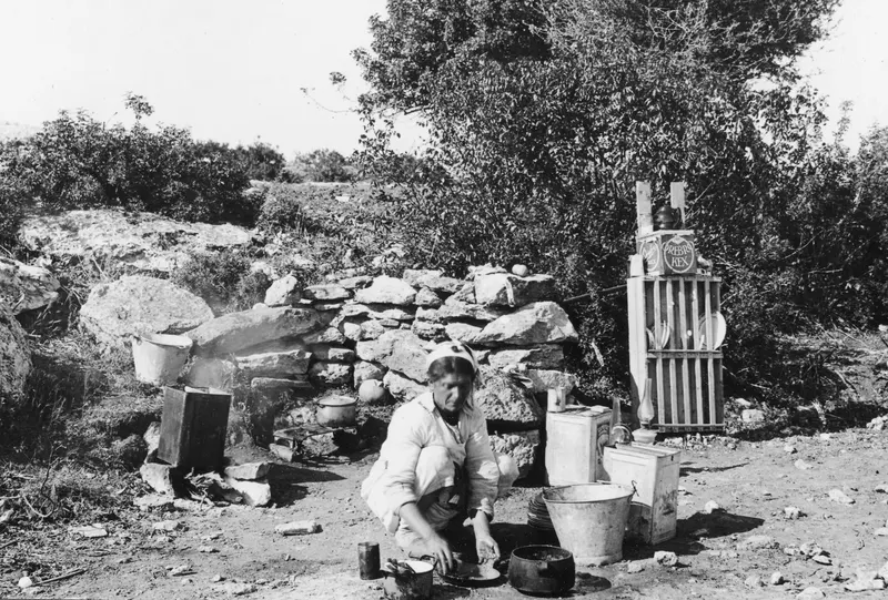 Eleni kneeling outdoors at Vouni, washing dishes beside a stone wall, with a dish rack and buckets nearby.