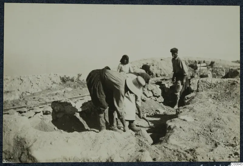 Workers bending over an excavation trench at Vouni, with a rail track along the edge.