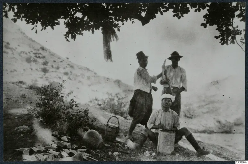 Three men at Vouni, one seated in a white cap and two standing behind him, one holding a hoe, with pottery on the ground.