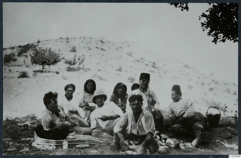 Nine people sit and rest on a striped mat by a hill at Vouni, with cups and a basket nearby.