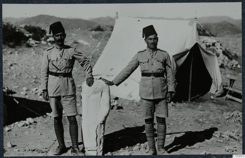 Two men in khaki uniform and fez caps beside a pale limestone statue, with a white tent behind them at Vouni