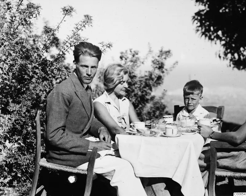 Three people at breakfast at a laid round table outdoors, with bushes behind them.