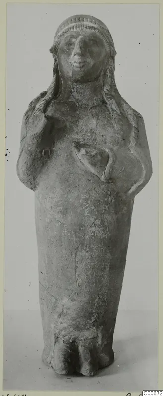 Standing terracotta figurine with arms bent up toward the chest and a band-patterned headdress, against a grey background