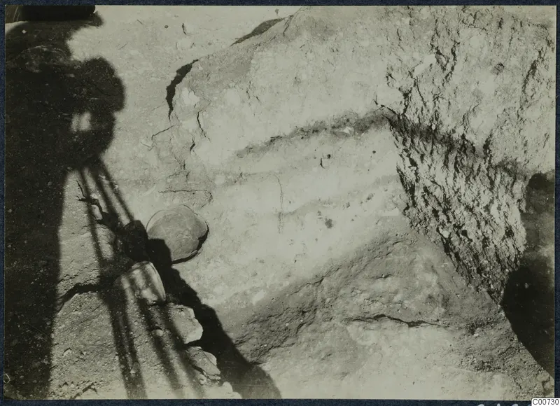 Excavation at Vouni palace: a layered earth face on the right, with a rounded vessel and stones in shadow on the left.