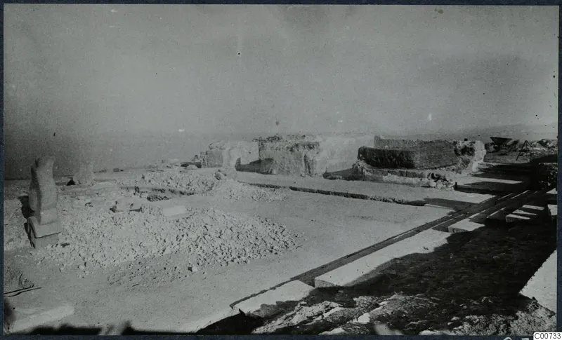 Excavated area at Vouni palace with rubble heaps, an upright stone, long stone slabs in the foreground and ruined walls behind.
