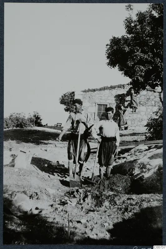 Two men leaning on spades beside a stone house under construction at Vouni