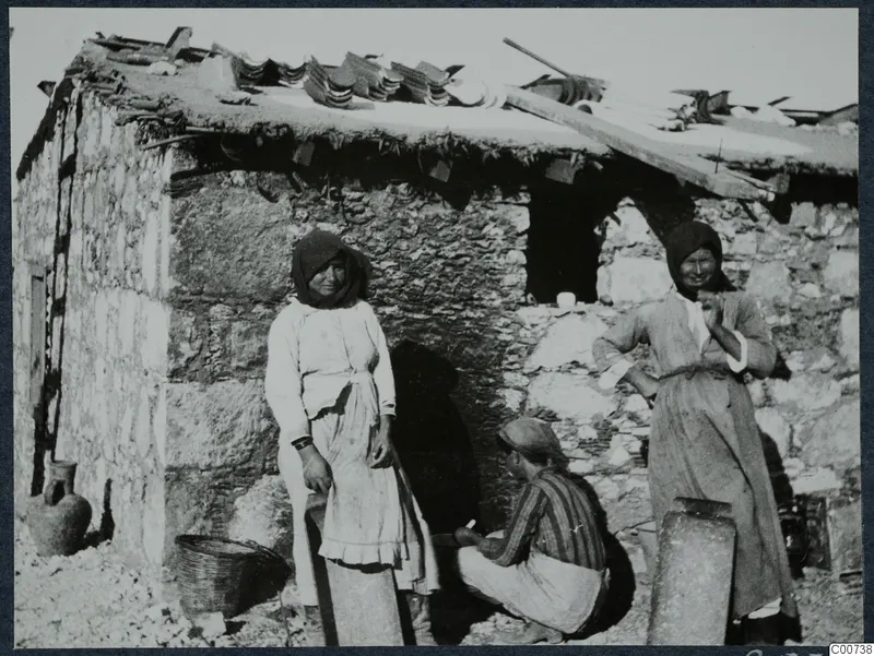 Two women and a crouching man beside a stone house with tiles and timbers on its roof at Vouni
