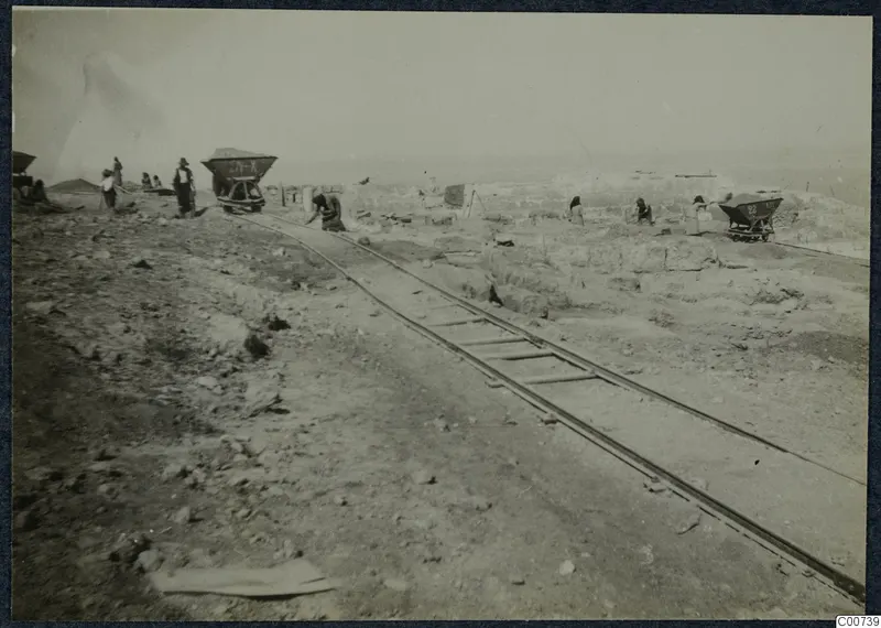 Excavation at Vouni with narrow rails, two tipping wagons and workers among low stone walls and rubble.