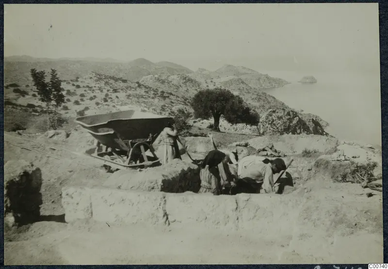 Four people digging with long-handled tools in an excavation at Vouni, with a wheelbarrow on rails and the sea beyond