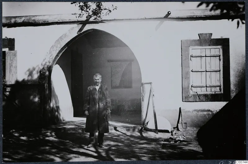 Man in a long coat standing outside the mukhtar's house in Milia, in front of a whitewashed arched doorway