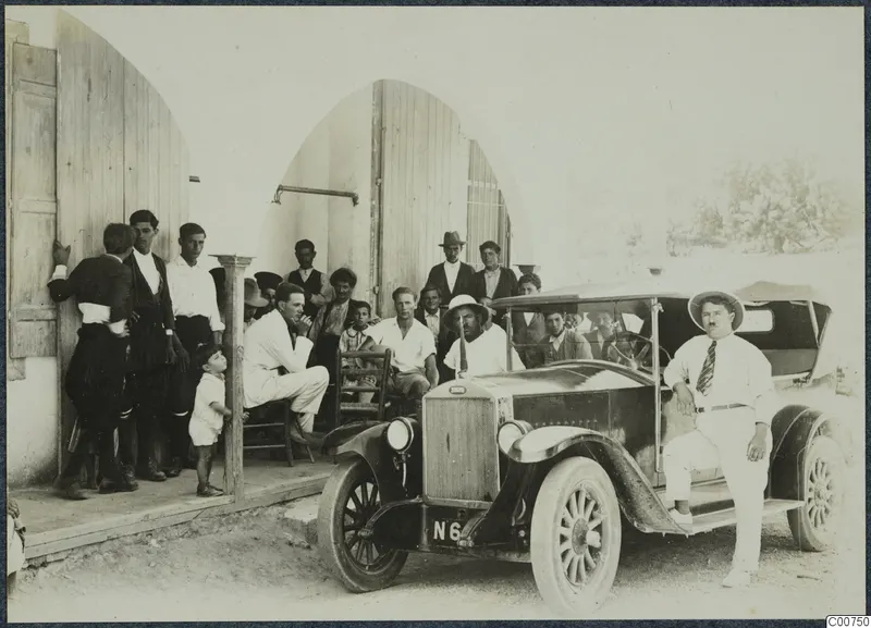 Group of men and a small child posing outside an arched doorway beside an open-top car with a tall radiator, in Pyrgos