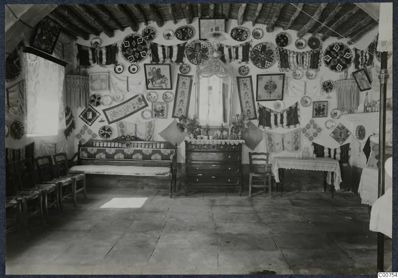 Interior of the mukhtar's best room at Milia, walls hung with embroidered textiles, framed pictures and decorated plates.