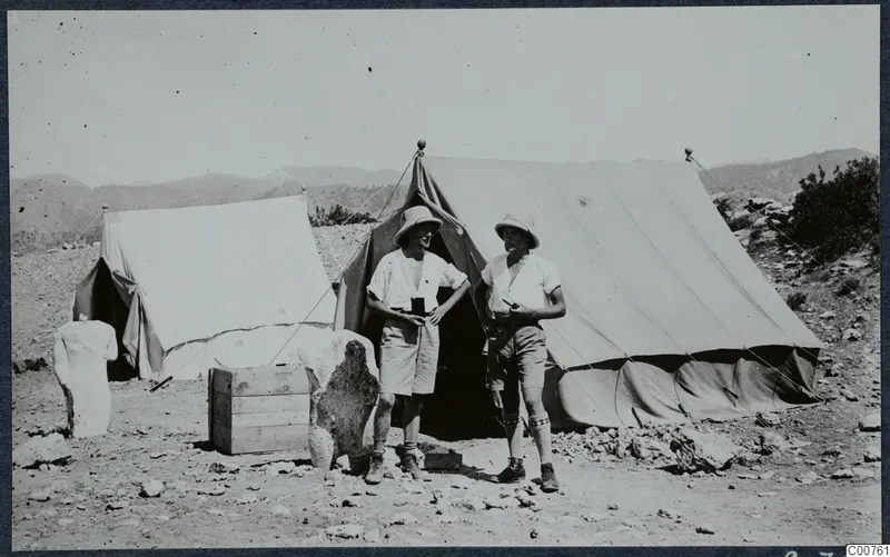 Two men in pith helmets stand outside large canvas tents at Vouni, with a stone sculpture fragment at left.