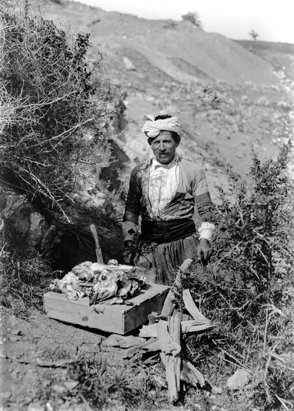 Man in a head cloth and sash stands beside a wooden box holding a butchered animal carcass, with firewood and scrub around him.