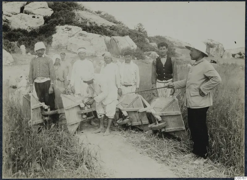 Workers with two wooden wheelbarrows on a grassy path at Oura, with a man in a hat standing to the right.