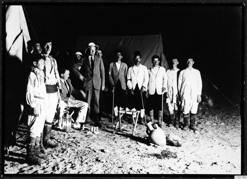 Men standing and seated outside dark tents on sand in low light, with a large two-handled amphora on the ground in front.