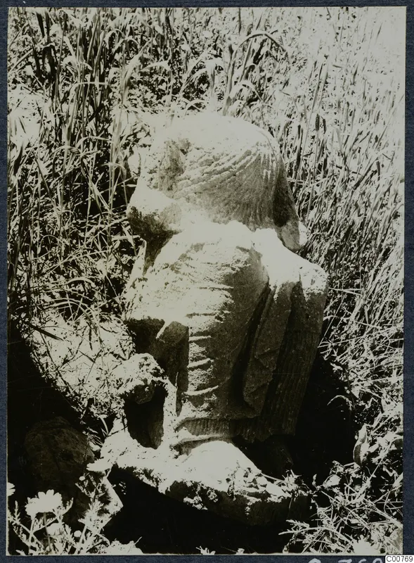 Weathered limestone statue of a figure, partly buried in tall grass at Oura.