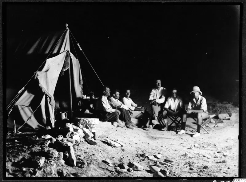 Six men seated together outdoors at night in front of a canvas tent, one in a pith helmet on the right.