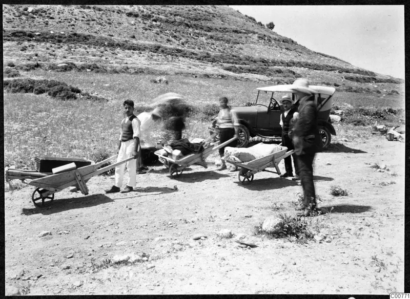 Men with three wheelbarrows loaded with bundles and boxes on a dry hillside, with a car behind them