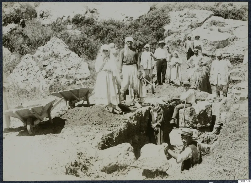Workers with shovels beside a trench and two wheelbarrows during excavation at Oura